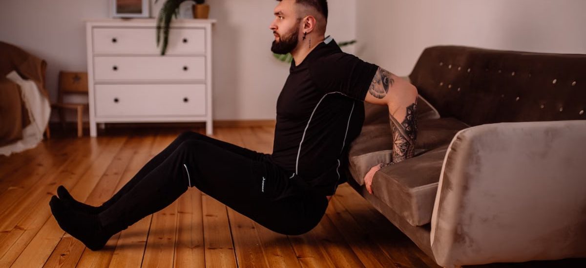 A bearded man in black fitness attire exercising at home, showcasing arm tattoos and a healthy lifestyle.