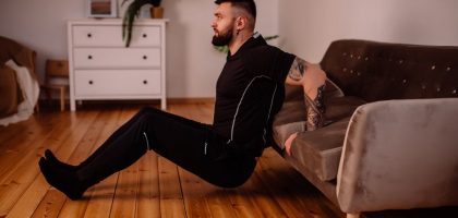 A bearded man in black fitness attire exercising at home, showcasing arm tattoos and a healthy lifestyle.